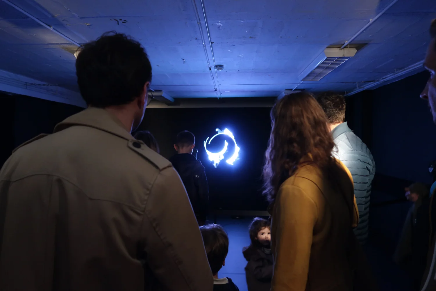 Vus de dos dans une salle obscure, des spectateurs de tous âges regardent l'anneau bleu suspendu devant eux.
