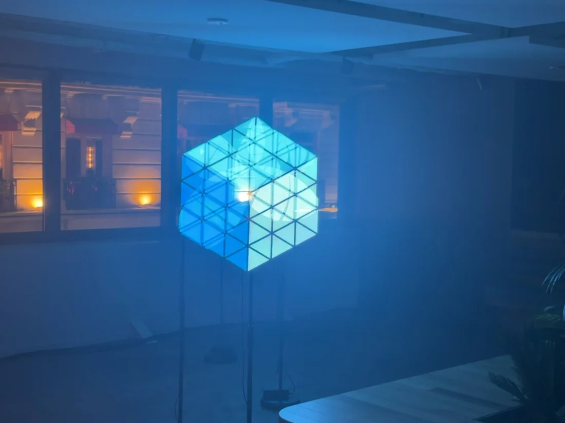 Dans un bureau plongé dans le bleu, quatre hélices recomposent un cube turquoise en fils de lumière, suspendu au milieu de la pièce.