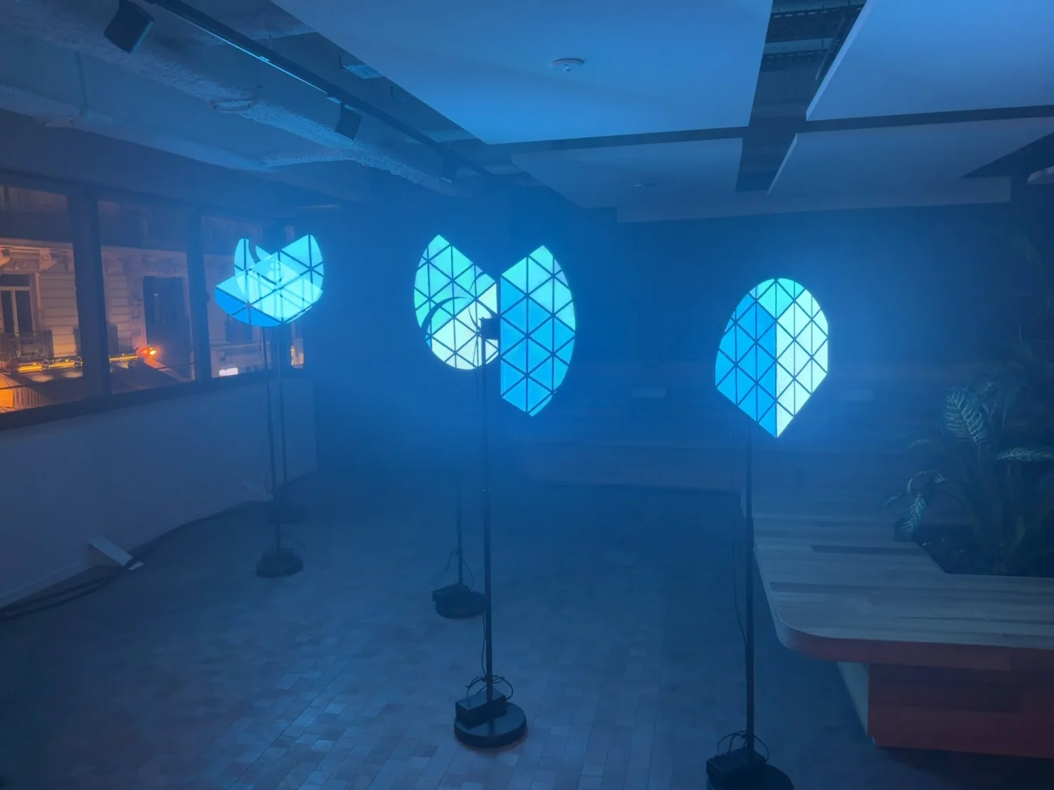 Quatre hélices sur pied dans un bureau bleuté, chacune portant un fragment lumineux qui ne se rassemble qu'à un endroit précis de la pièce.