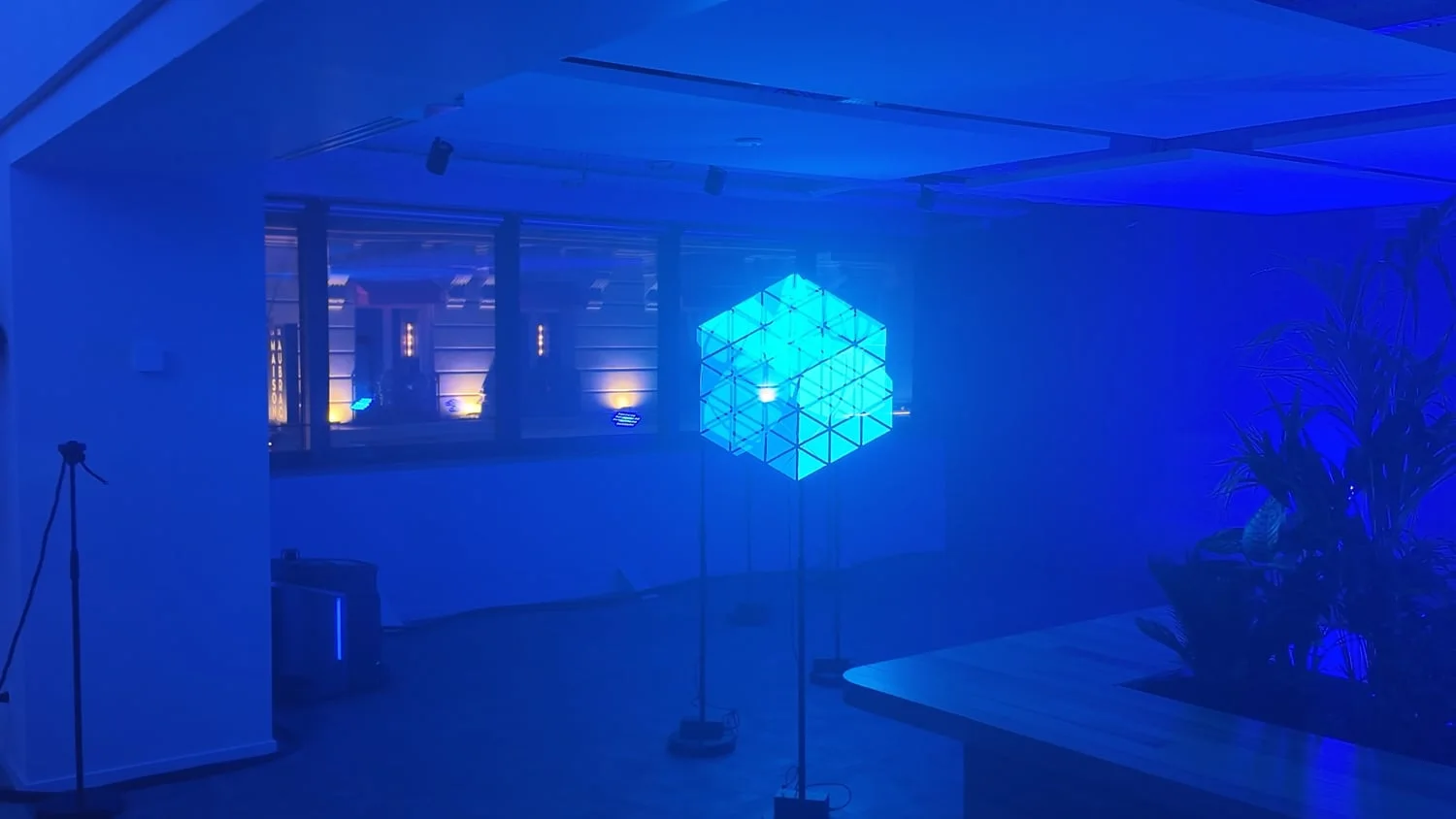 Image extraite de la vidéo : dans une salle baignée de bleu, plusieurs hélices sur pied recomposent un cube lumineux en fils turquoise, suspendu au milieu de la pièce.