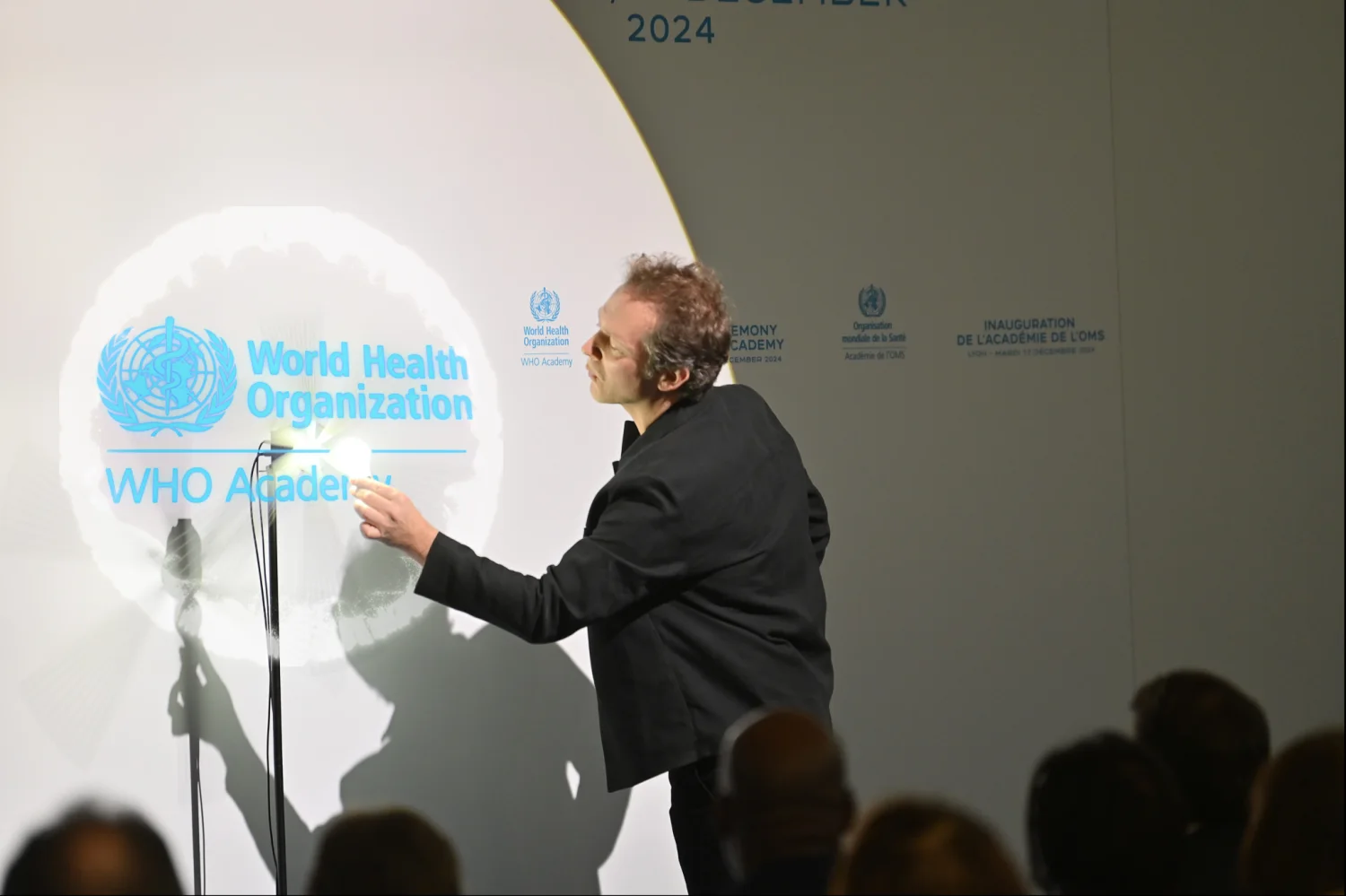 Inauguration de l&rsquo;Académie de l&rsquo;OMS à Lyon : le magicien fait apparaître le logo de l&rsquo;Organisation mondiale de la santé dans l&rsquo;hélice, devant le public assis.