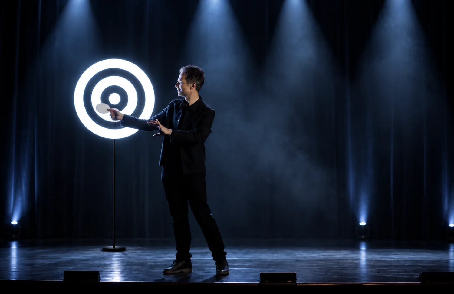 Sur une scène de théâtre, le magicien tend la main vers l&rsquo;hélice sur pied : une cible lumineuse concentrique tourne dans la fumée.