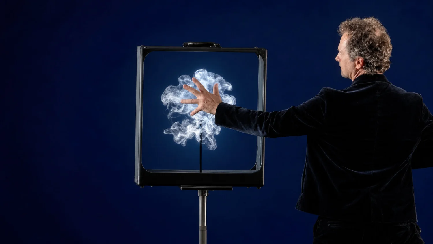 Sur fond bleu nuit, le magicien de dos pose la main sur le cadre de la valise posée sur pied : une volute de fumée blanche tourne à l&rsquo;intérieur.