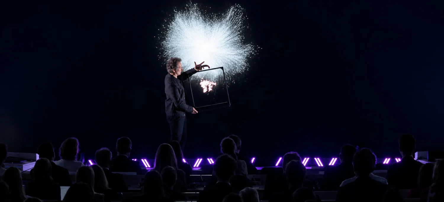 Sur scène, face au public assis, Romain Lalire tient le cadre de la valise à bout de bras : une gerbe de particules blanches explose au-dessus, prolongeant l&rsquo;hologramme hors du cadre.